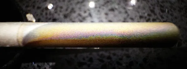 Close-up of a metal electrode rod with a shimmering band of gold, pink and green colours across its surface