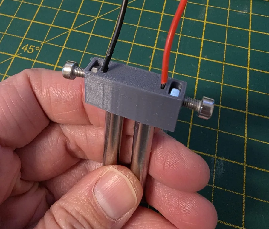 A 3D-printed grey block with two screws, holding a pair of stainless electrodes with a red and a black wire, held in a hand over a cutting mat