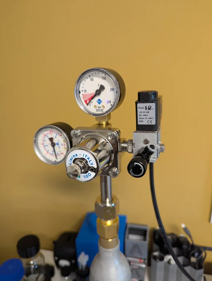 A CO₂ regulator with two pressure gauges on a SodaStream cylinder, with a small silver solenoid valve with a black coil fitted to its outlet