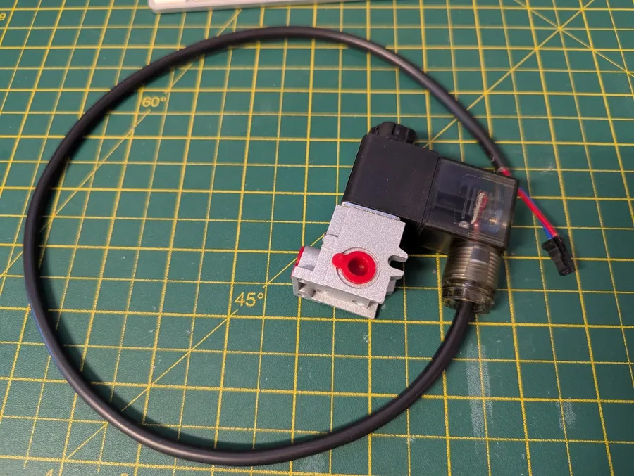 A small silver aluminium solenoid valve with a black coil, a transparent DIN plug and a loop of black cable, lying on a green cutting mat